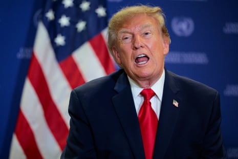 Donald Trump speaks animatedly in a dark suit and red tie in front of an American flag